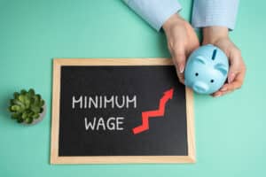 chalkboard with the words minimum wage and an upward arrow pointing towards hands holding a piggy bank